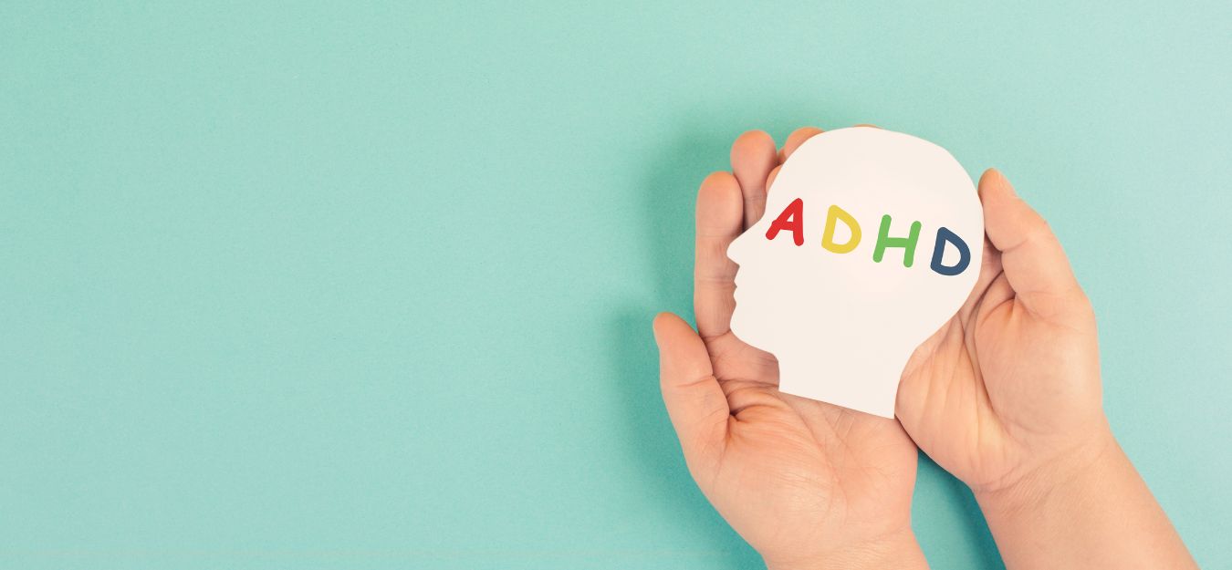 Two hands holding a paper cut-out of a head that says ADHD in rainbow letters. the background is light blue.