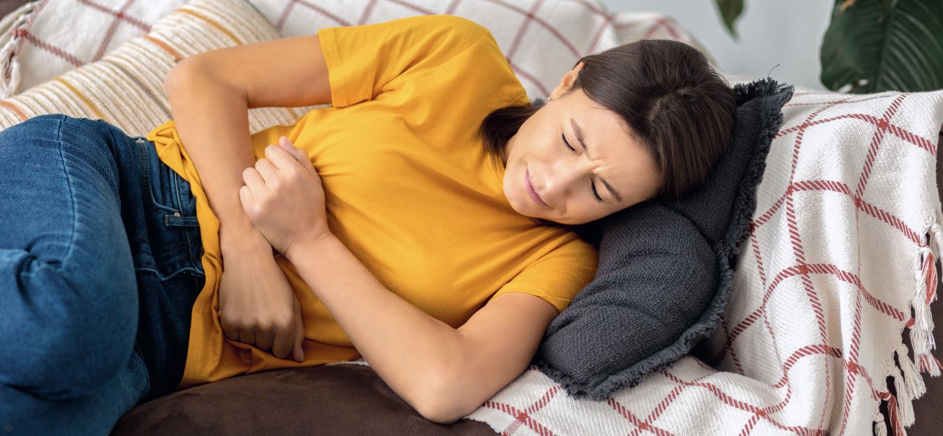 A woman in a yellow sweater laying on a couch, holding her stomach as if in pain.