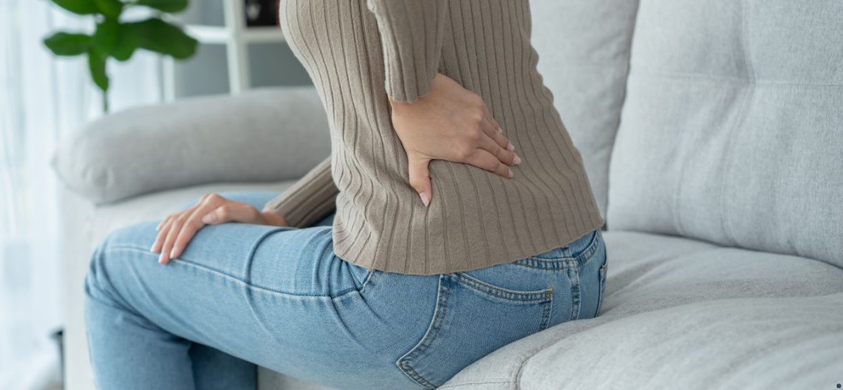 A woman sitting on a couch, holding a hand over her lower back as if experiencing pain.