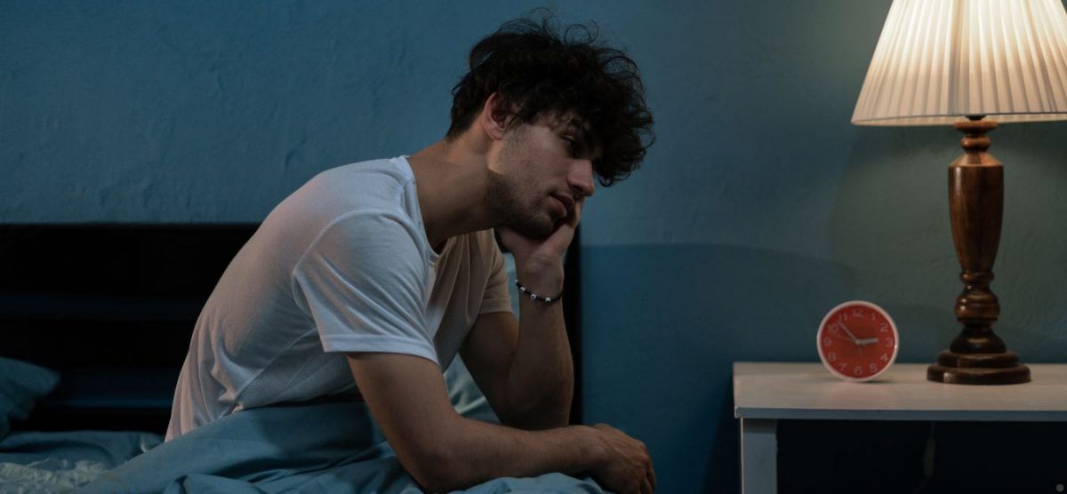 A man sitting up in bed, unable to sleep. There is a lamp and alarm clock on his nightstand.