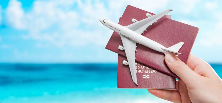 A hand holding two passports and a toy plane in front of the ocean.